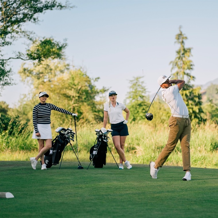 Three golfers showcasing stylish golf wear on a green fairway. A man in a white polo shirt and khaki pants is mid-swing, while two women, one in a striped shirt and white skirt and the other in a white polo and navy shorts, watch with their golf bags beside them.