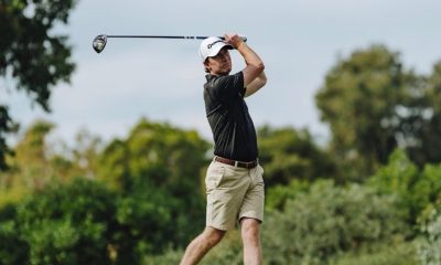 A male golfer in trendy golf wear, consisting of a black polo shirt and beige shorts, follows through on his swing on a lush green golf course, with trees and bushes in the background.